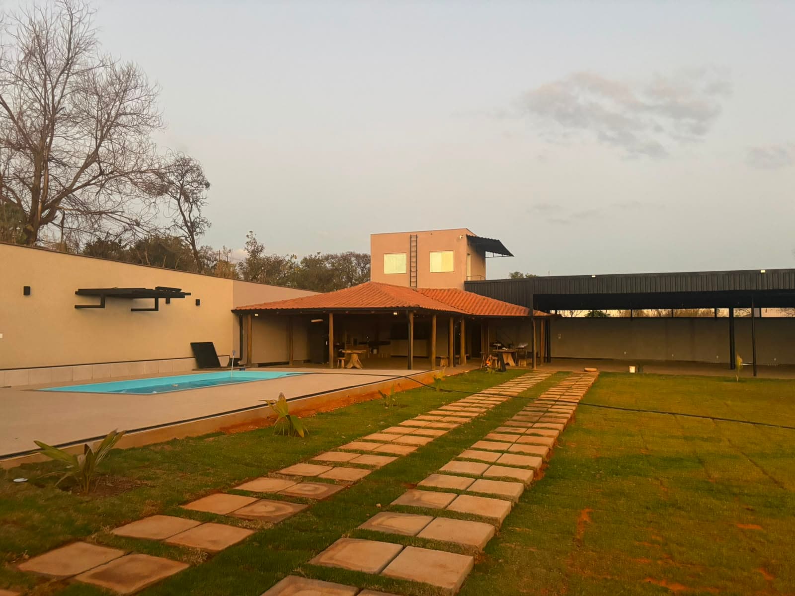 Vista do gramado ao entardecer, com a piscina, o quiosque gourmet e o salão coberto ao fundo.